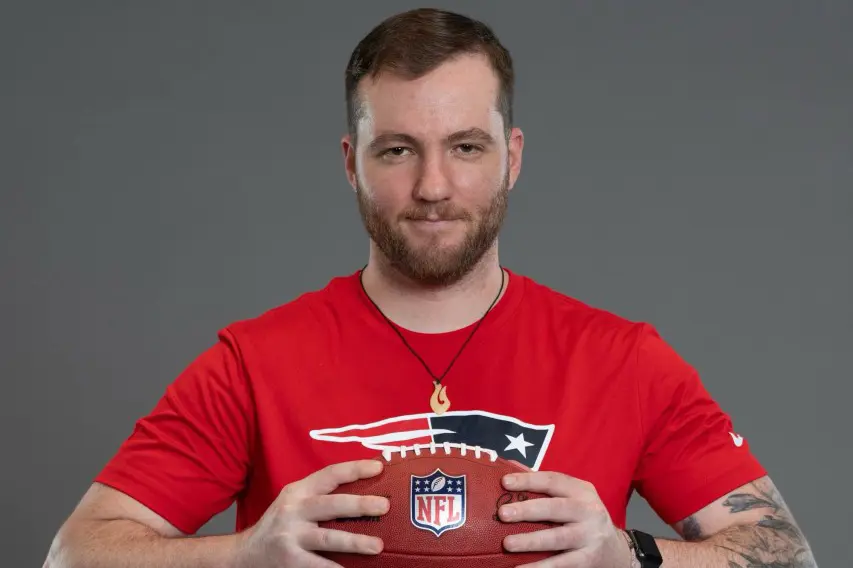 A young man with a beard poses for a photo while wearing a red Patriots T-shirt and holding and NFL football.
