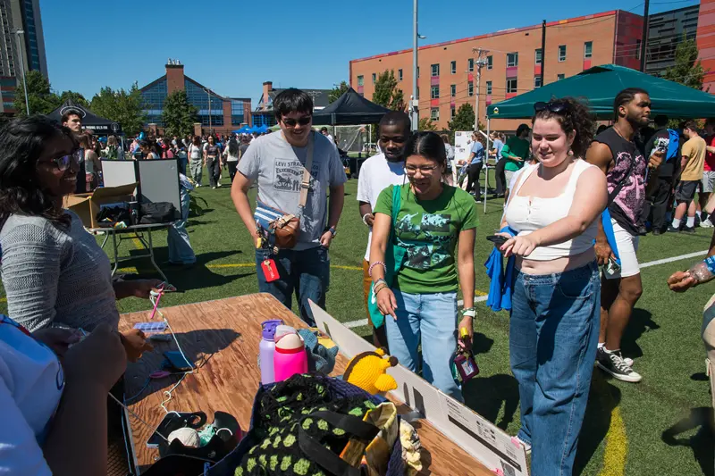 Four students approach at table tended by another student with giveaway on it at outdoor fair.