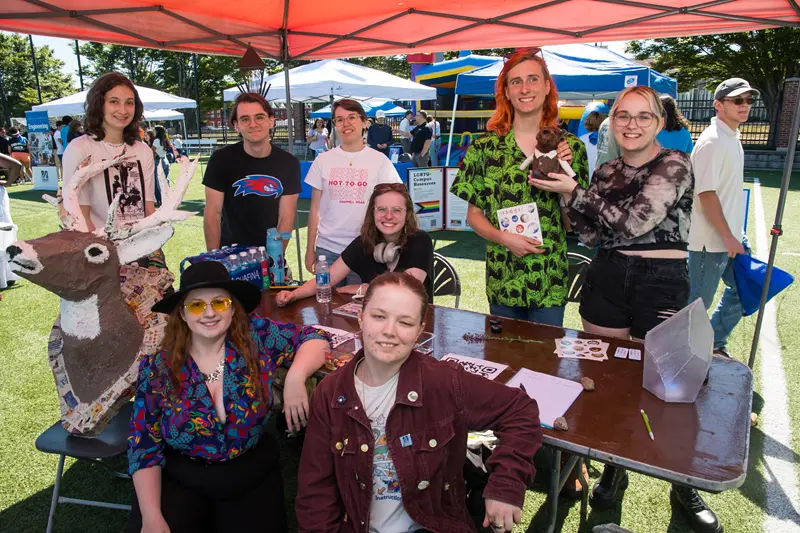 Students gather around table with paper mache deer and LGBTQ+ Campus Resources poster. One student wearing shirt that repeats Hot-to-go
