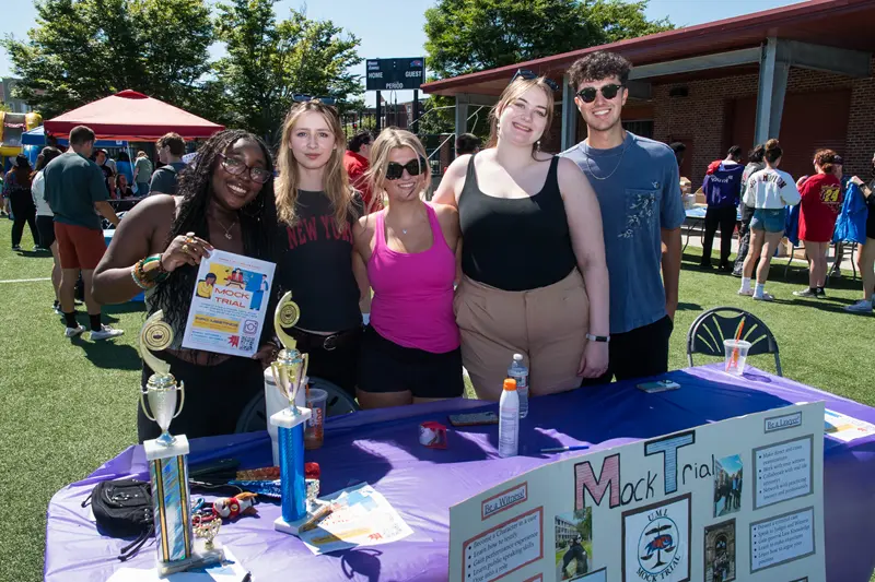 4 young women and a young man standing at the Mock Trial table with poster on front of table and one student holding a Mock Trial flyer