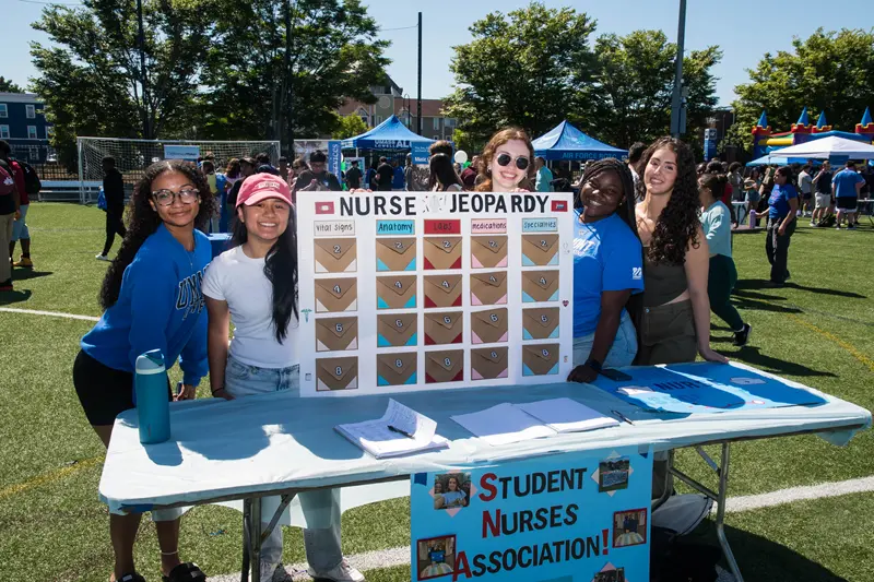 Five young women gather around a poster that says Nurse Jeopardy with a Student Nurses Association poster hanging from table.