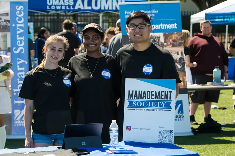 Three student standing behind a Management Society poster with Library Services and UMass Lowell Alumni signs in background