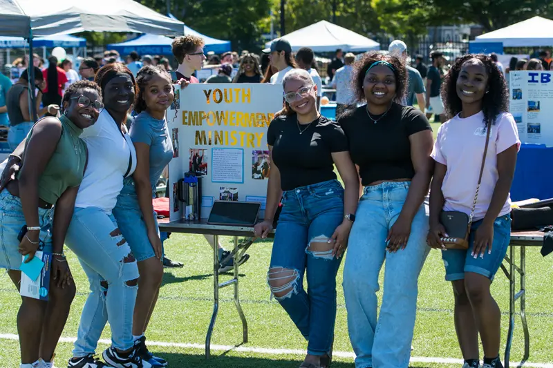 Six young women standing in front of a Youth Empowering Ministry poster