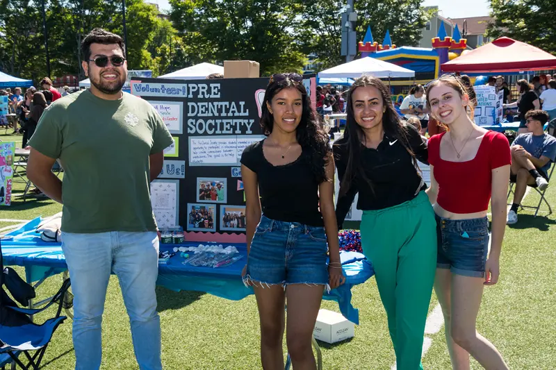 A young man and three young women in front of poster with Pre-Dental Society info on it