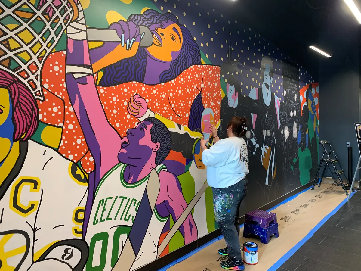 A woman paints a mural that has people painted in bright colors.