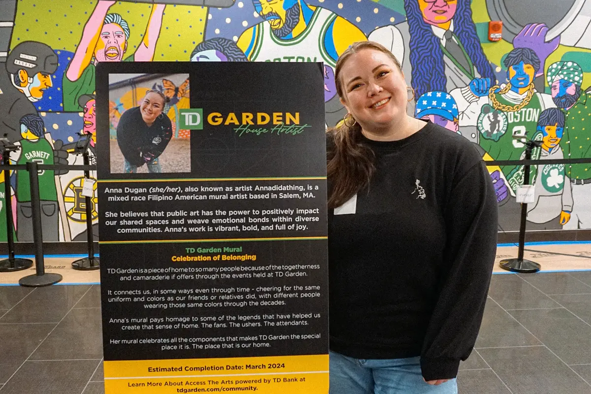 A woman in a black shirt poses with a sign in front of a mural.