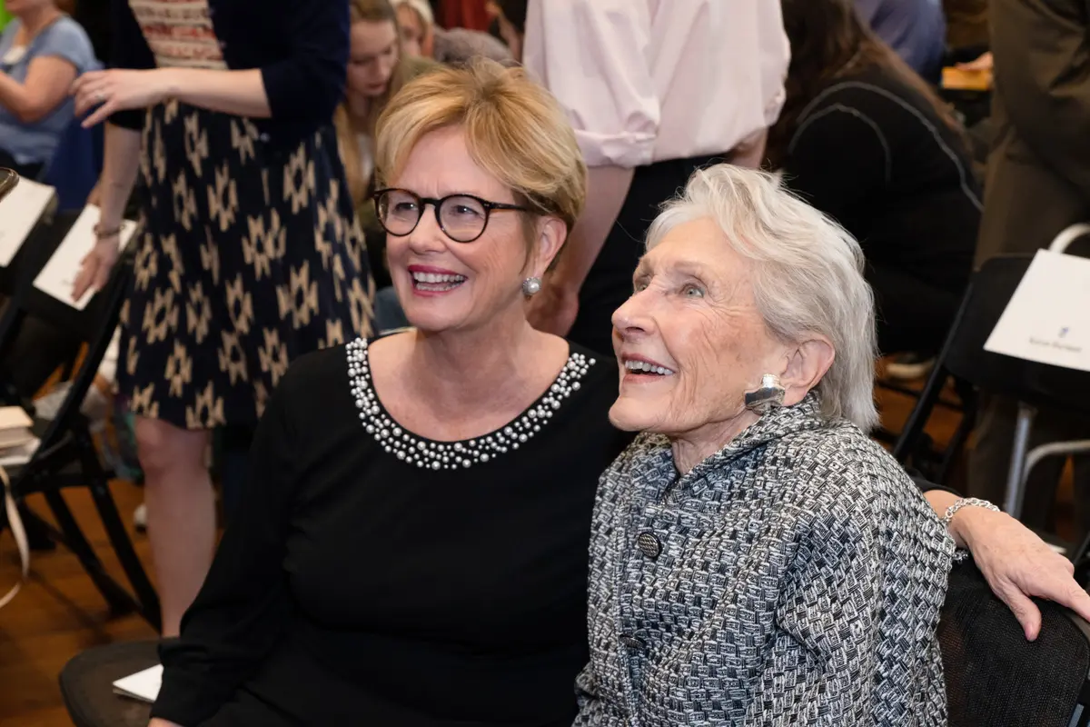 Two women smile while seated next to each other, looking up at someone.