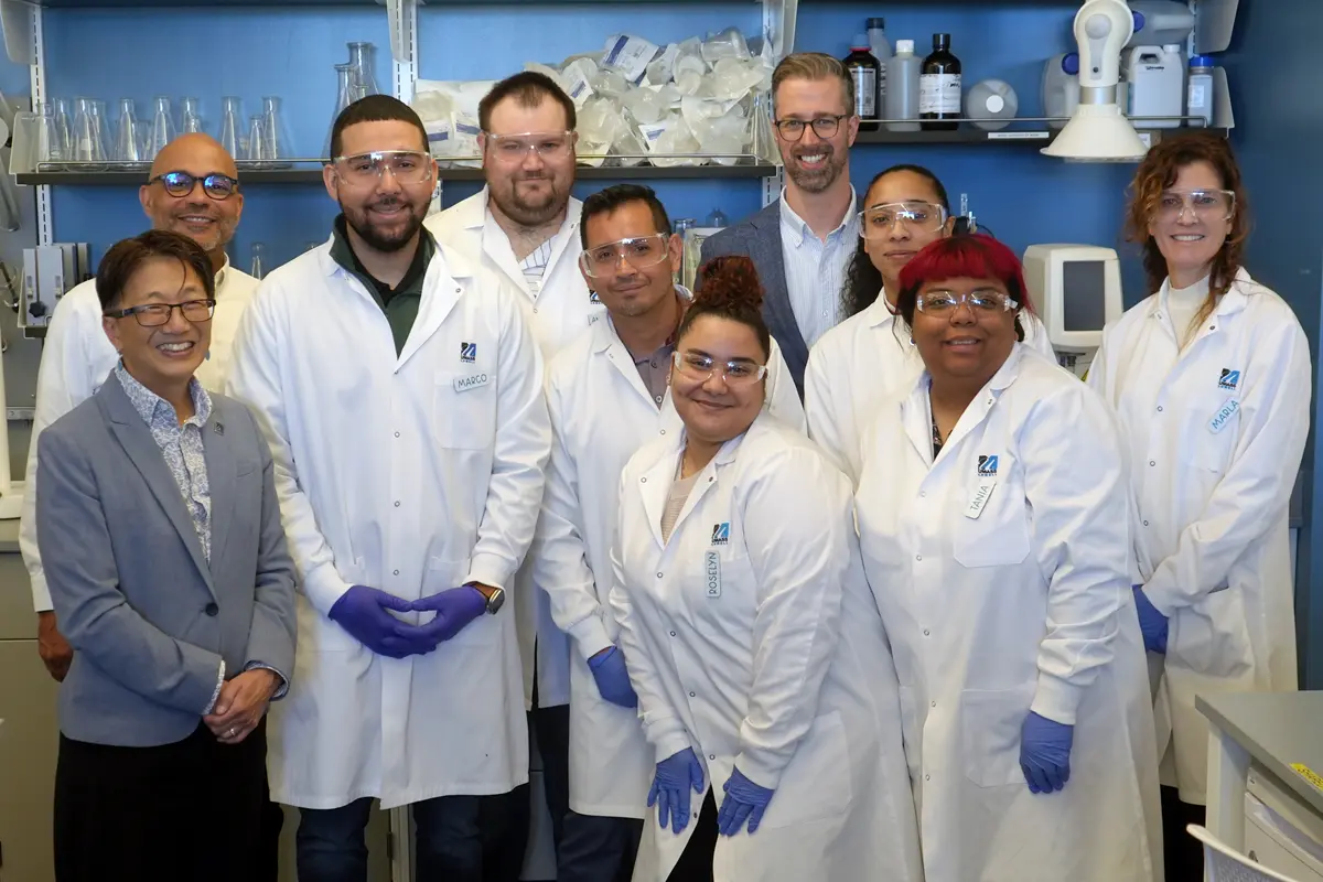 10 people stand in a lab, wearing glasses and lab coats.
