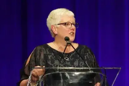 A woman with short hair and glasses speaks at a glass podium in front of a blue backdrop.