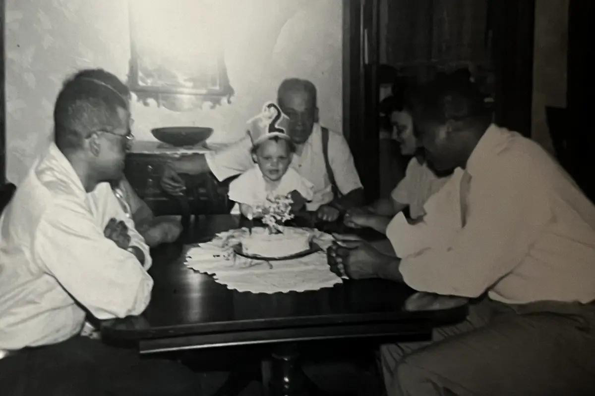 A group of people sit around a table while a little girl with a 2 hat looks at a birthday cake.