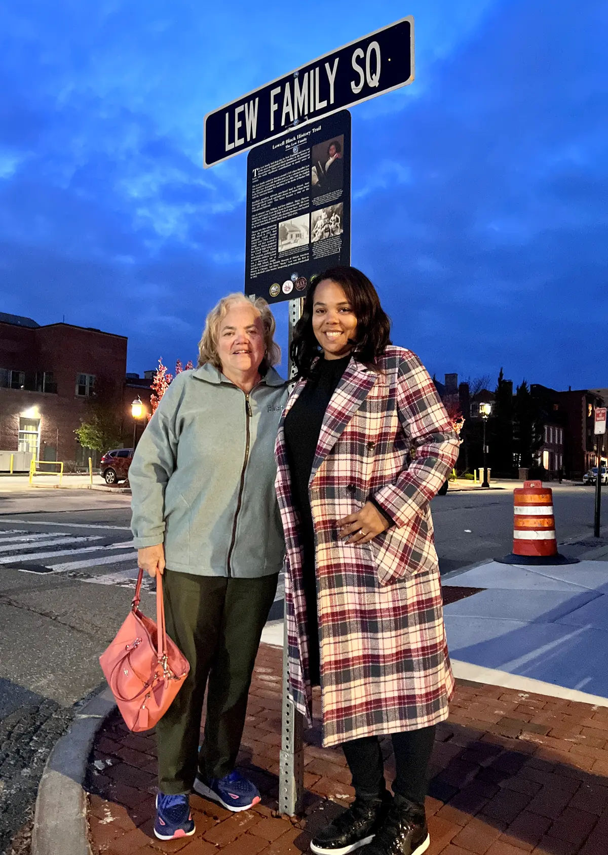 Two women pose for a photo in front of a street sign that says Lew Family Square