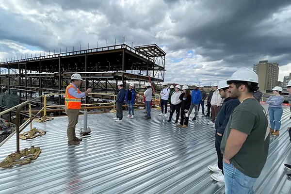 A man in an orange vest and hard hat gestures while talking to a group of students