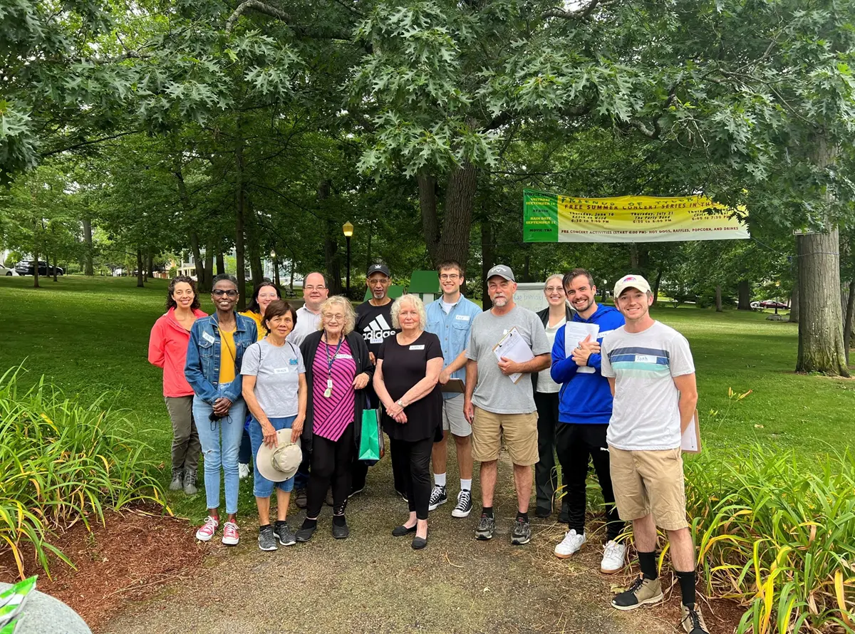 A group of 13 people standing in front of a line of trees in the back at a park.