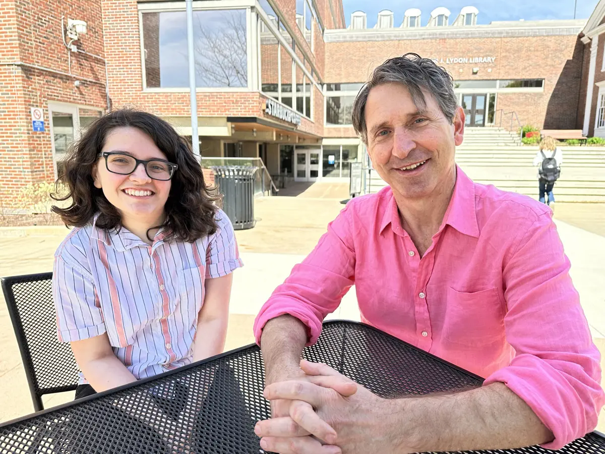 A college student with glasses poses for a photo while sitting at an outdoor table with a professor wearing a pink shirt.