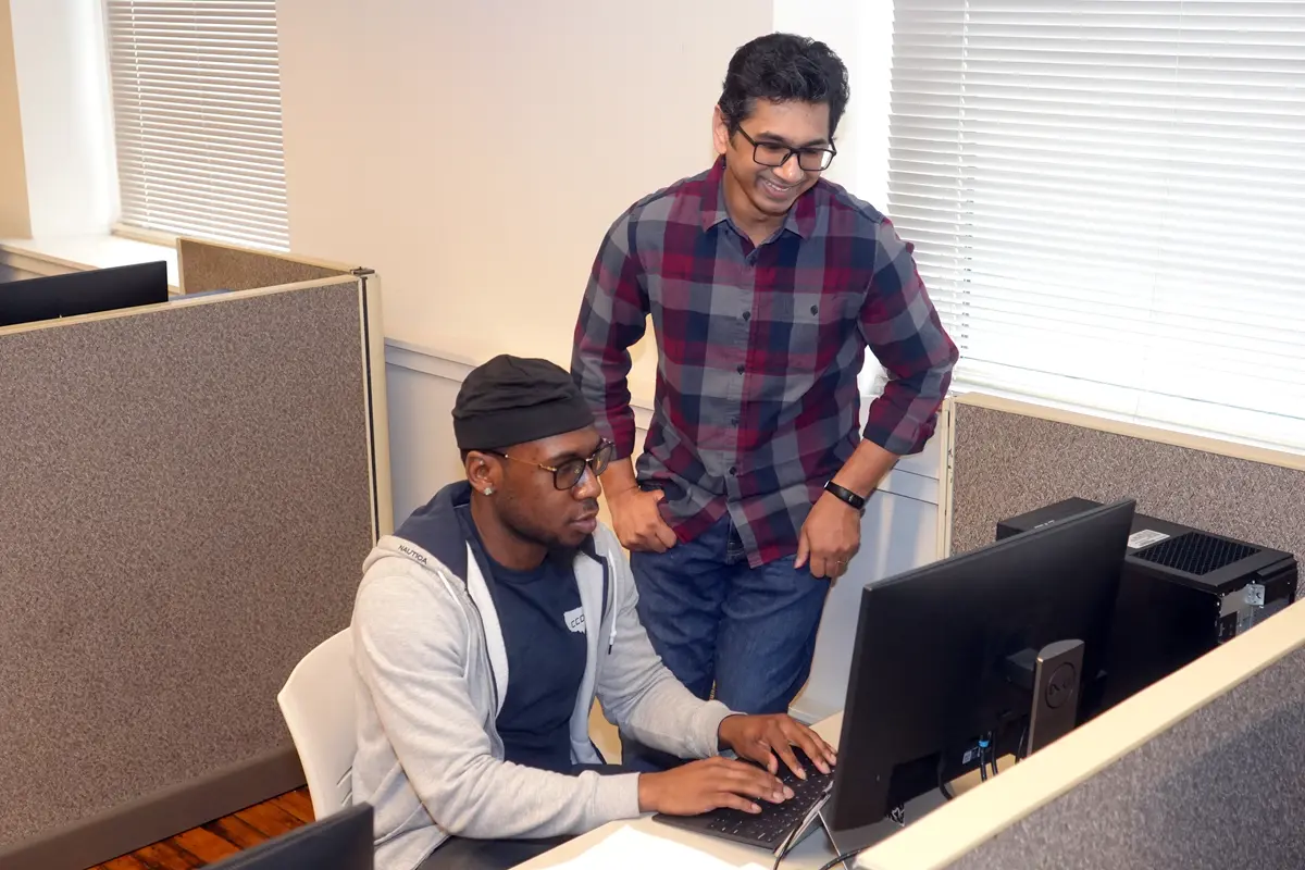 A seated young man types on a computer keyboard while a standing man looks on.