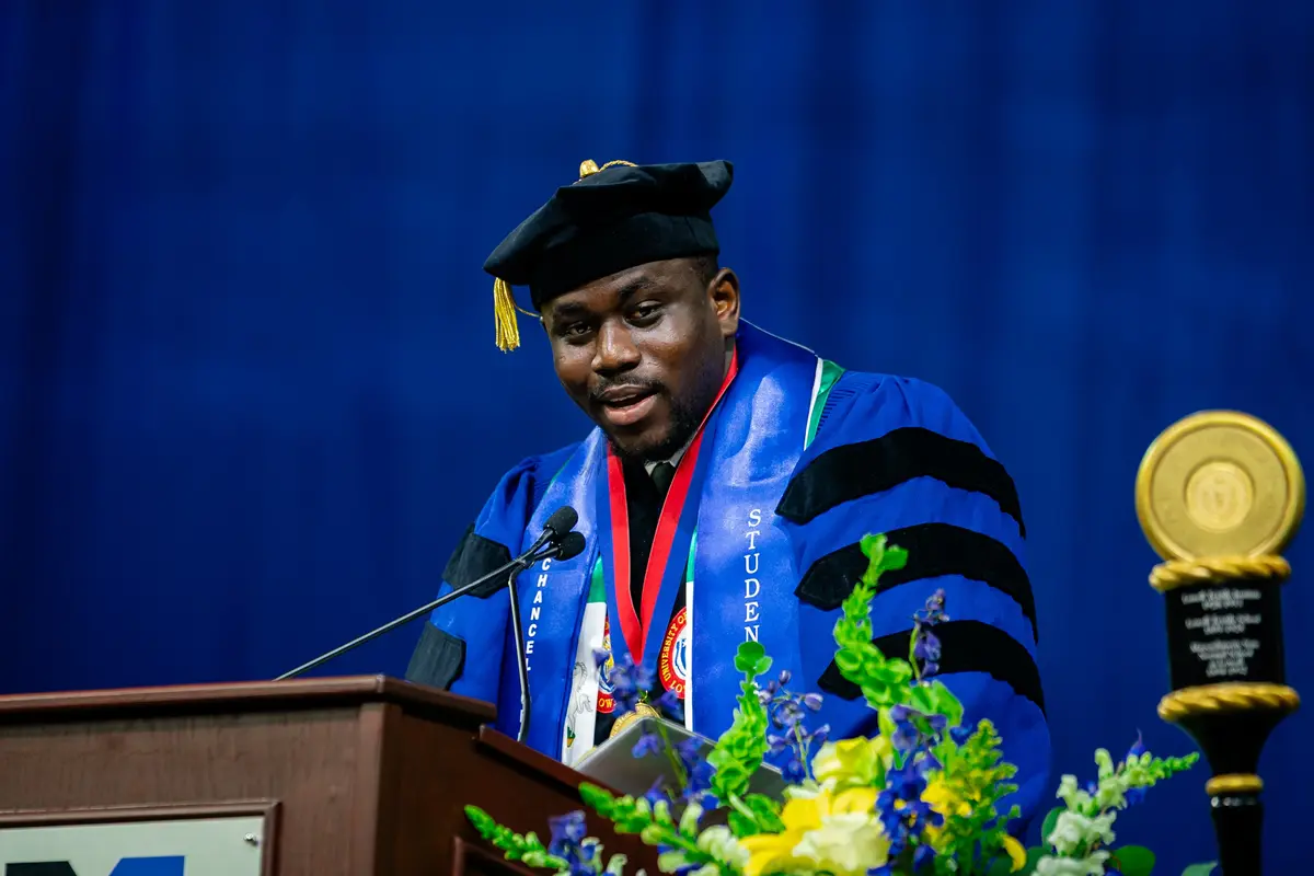 A student wearing academic regalia speaks at a podium during a commencement ceremony at UMass Lowell