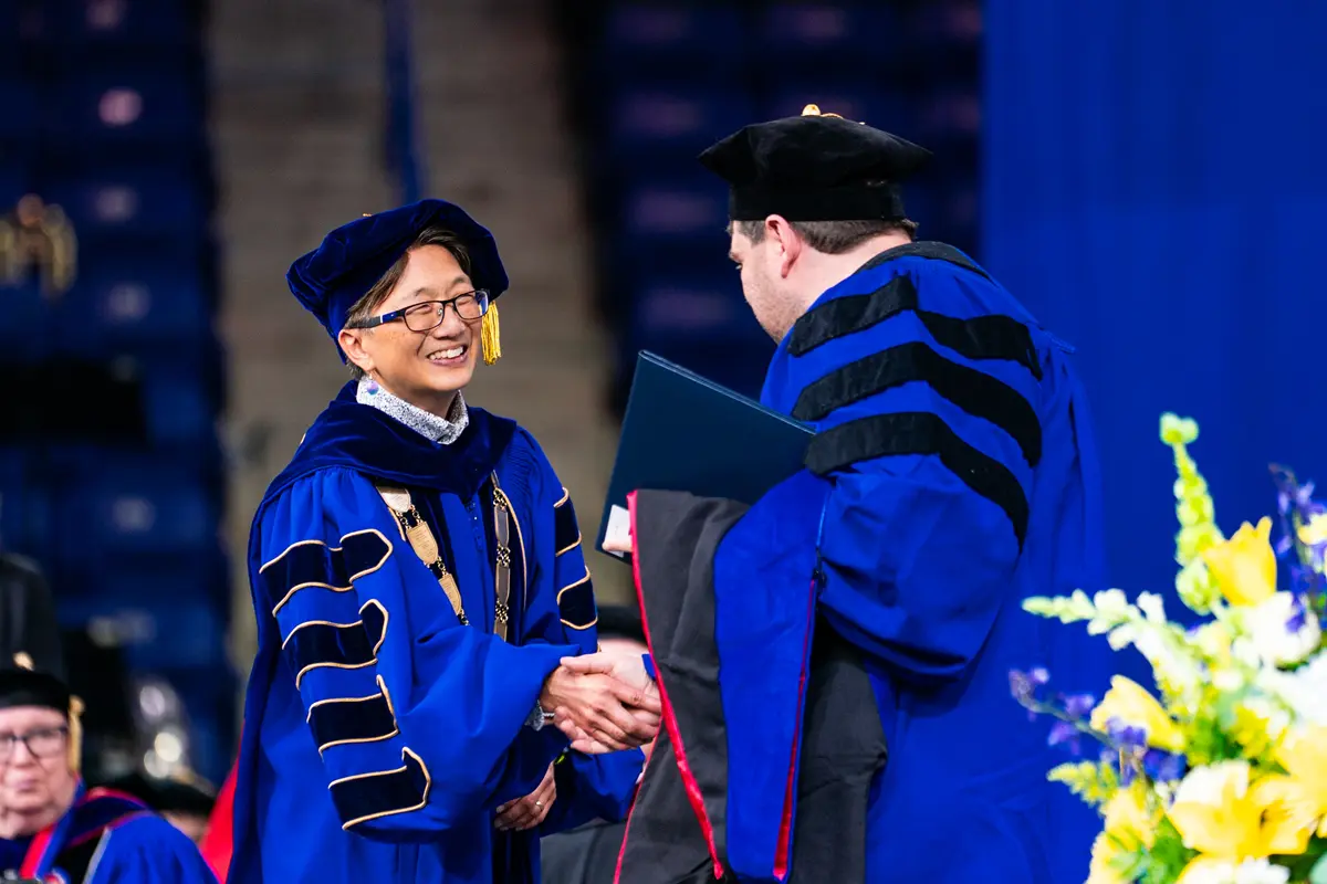 A man and woman wearing academic regalia shake hands during a ceremony on stage