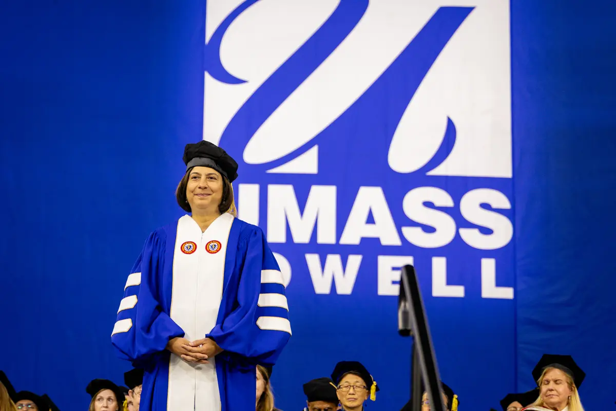 A woman wearing a blue and white UMass Lowell academic robe stands on stage with her fingers interlaced