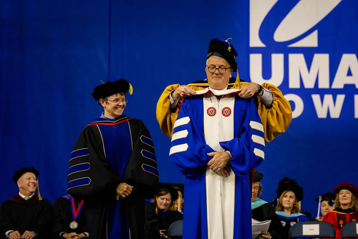 An honorary degree awardee is hooded by someone standing behind him as a woman dressed in academic regalia looks on