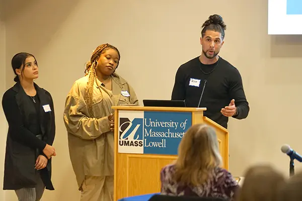 A man gestures while speaking at a podium while two women look on