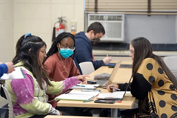 A professor in a polka dot top sits at a table and speaks with two female students