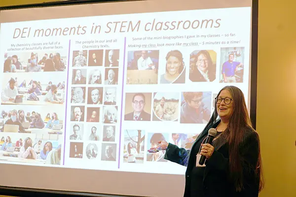 A woman wearing glasses and holding a microphone makes a presentation in front of a project screen