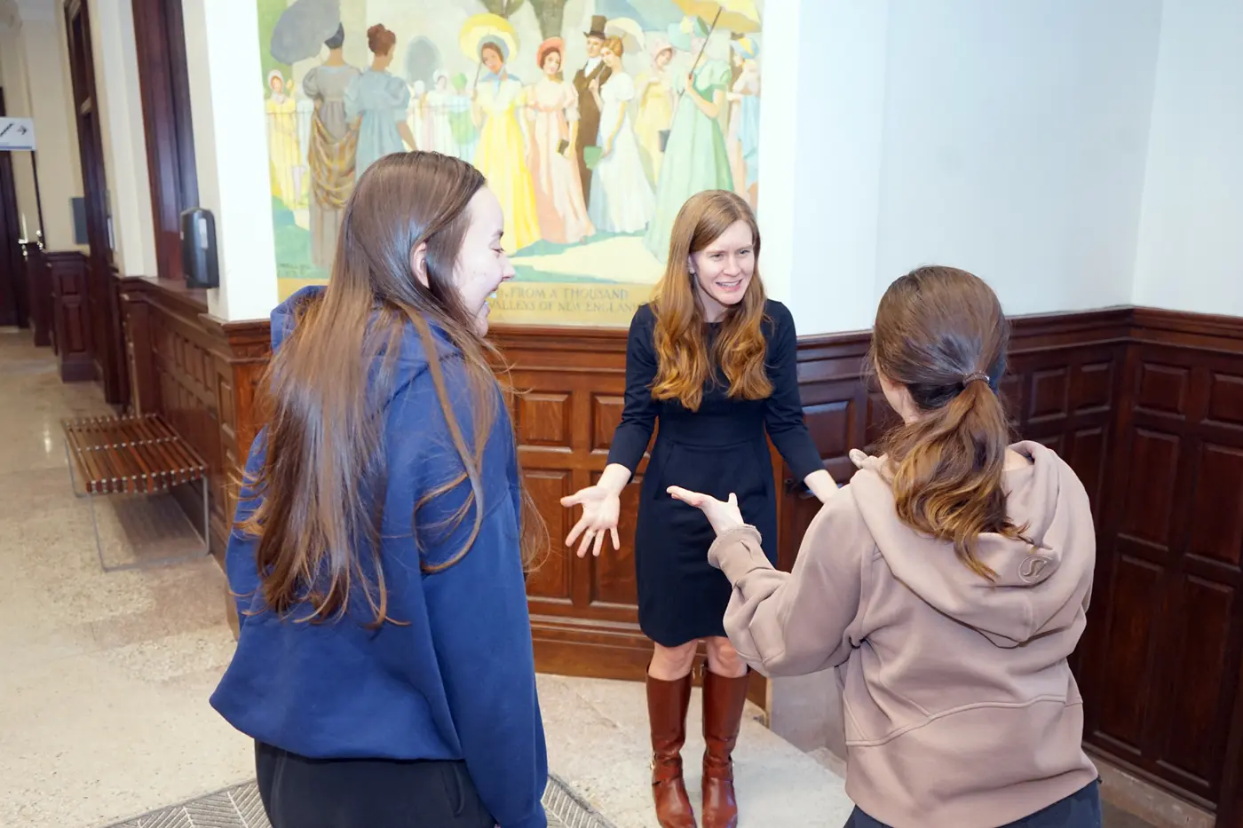 A woman in a dark dress and boots talks with two college students in the hallway of a building.