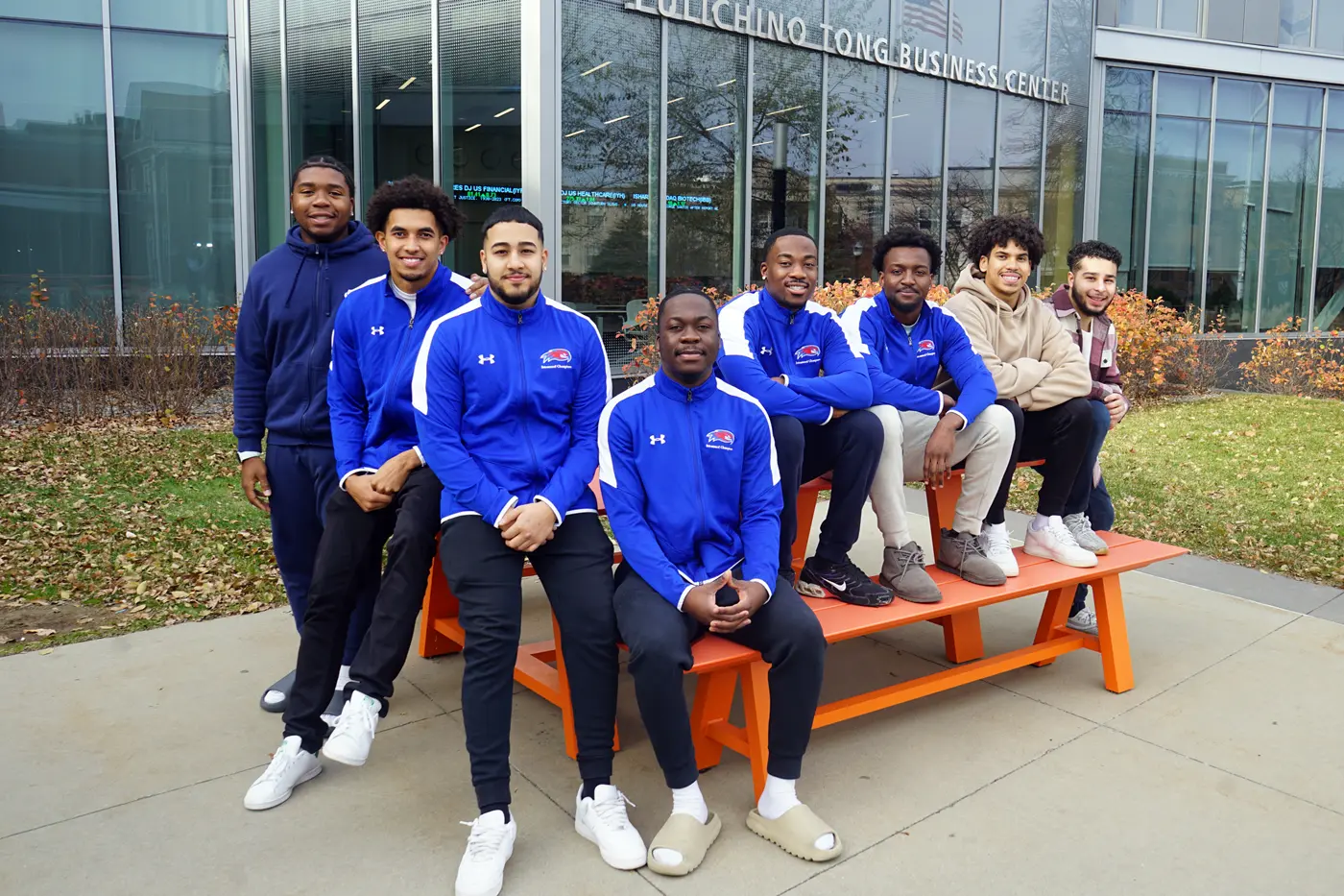 Eight people pose for a group photo while sitting on a picnic table outside of a building.