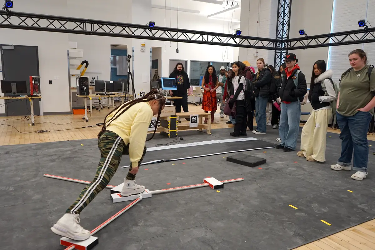 A young woman stretches her leg on a sliding board while a group of people look on in a robotics lab.