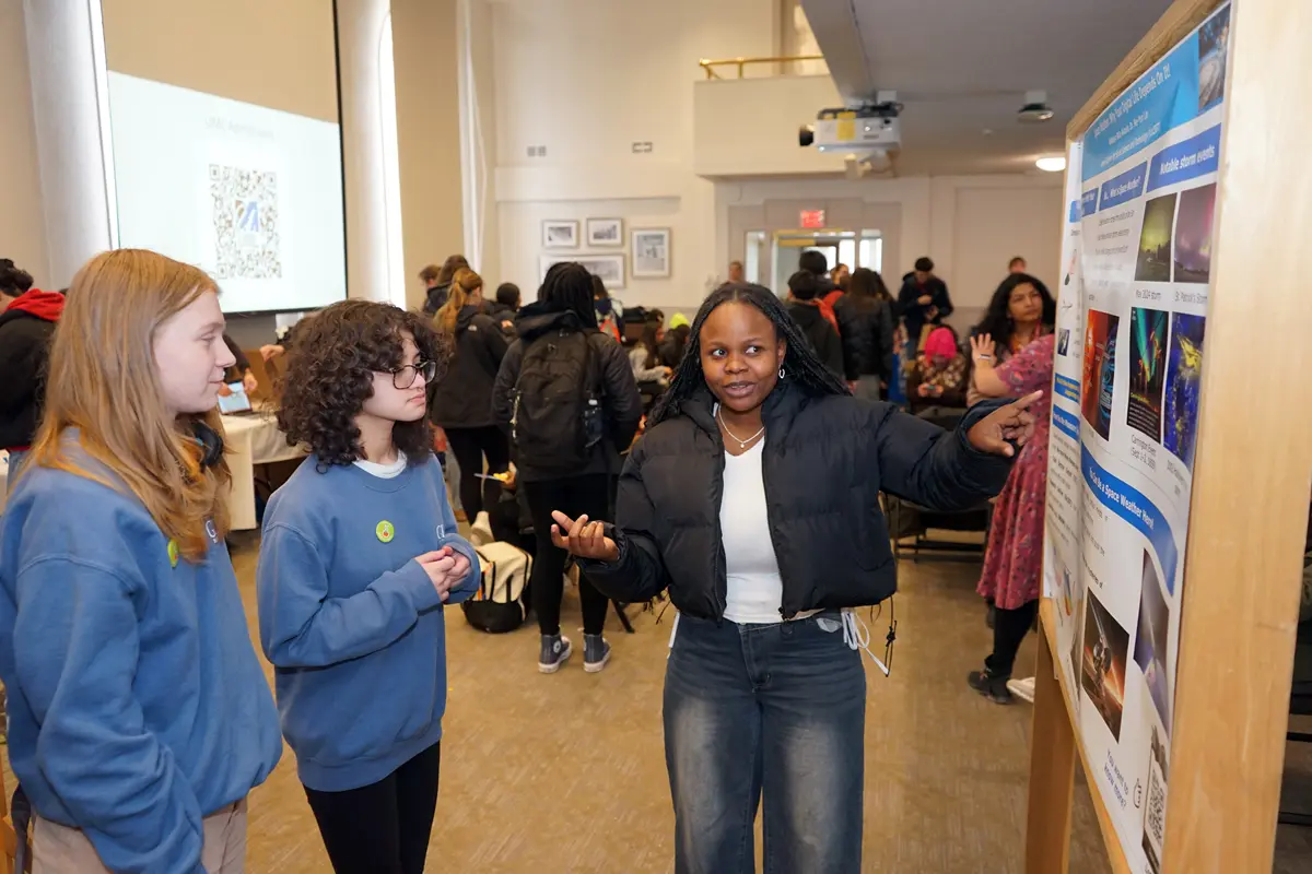 A young woman points to a research poster while talking to two high school students in a large room.