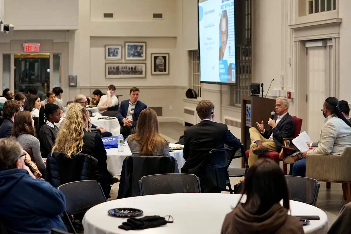 A seated man holds a microphone and talks to a room full of college students.