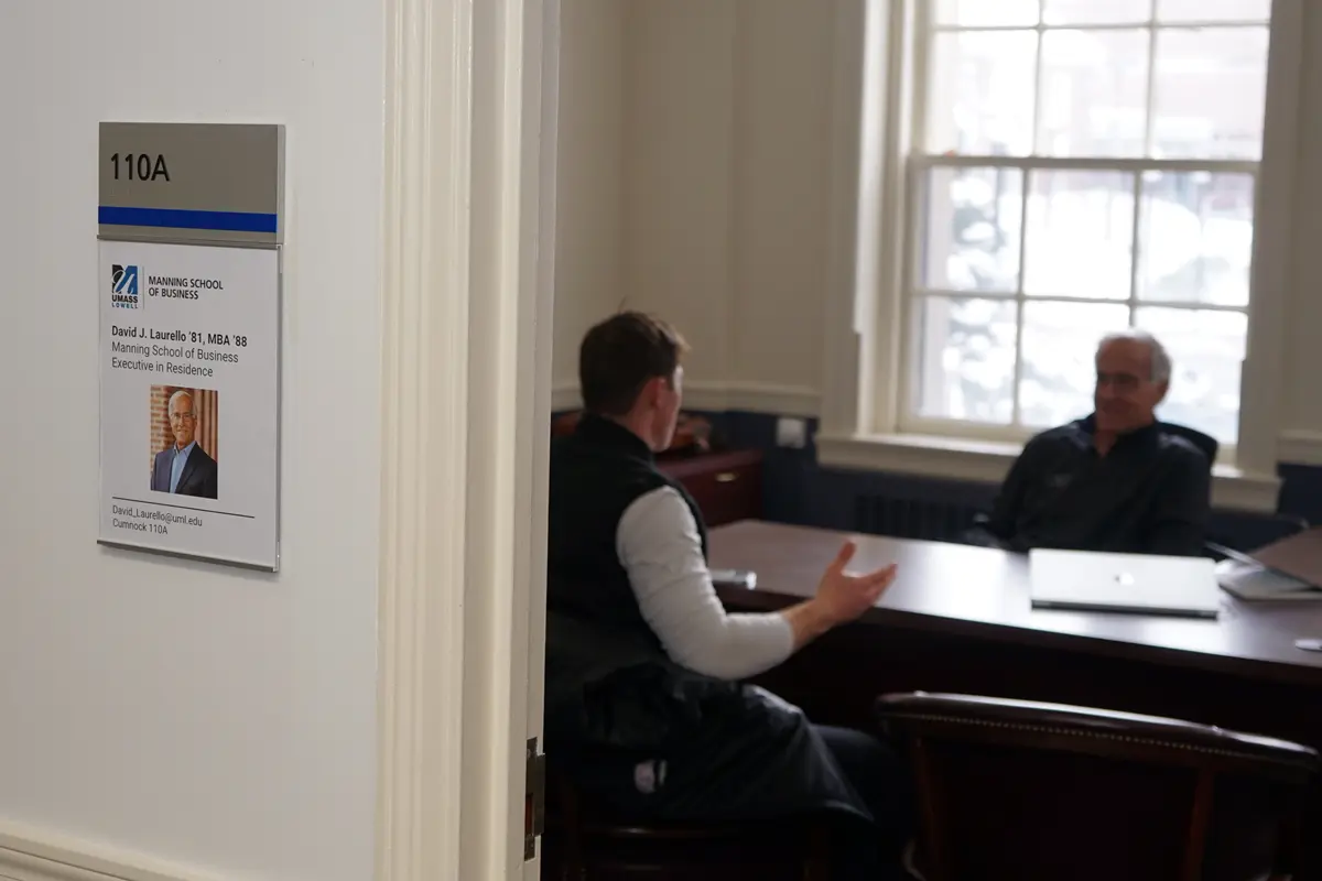 Two men can be seen through an open doorway talking while seated at a desk.