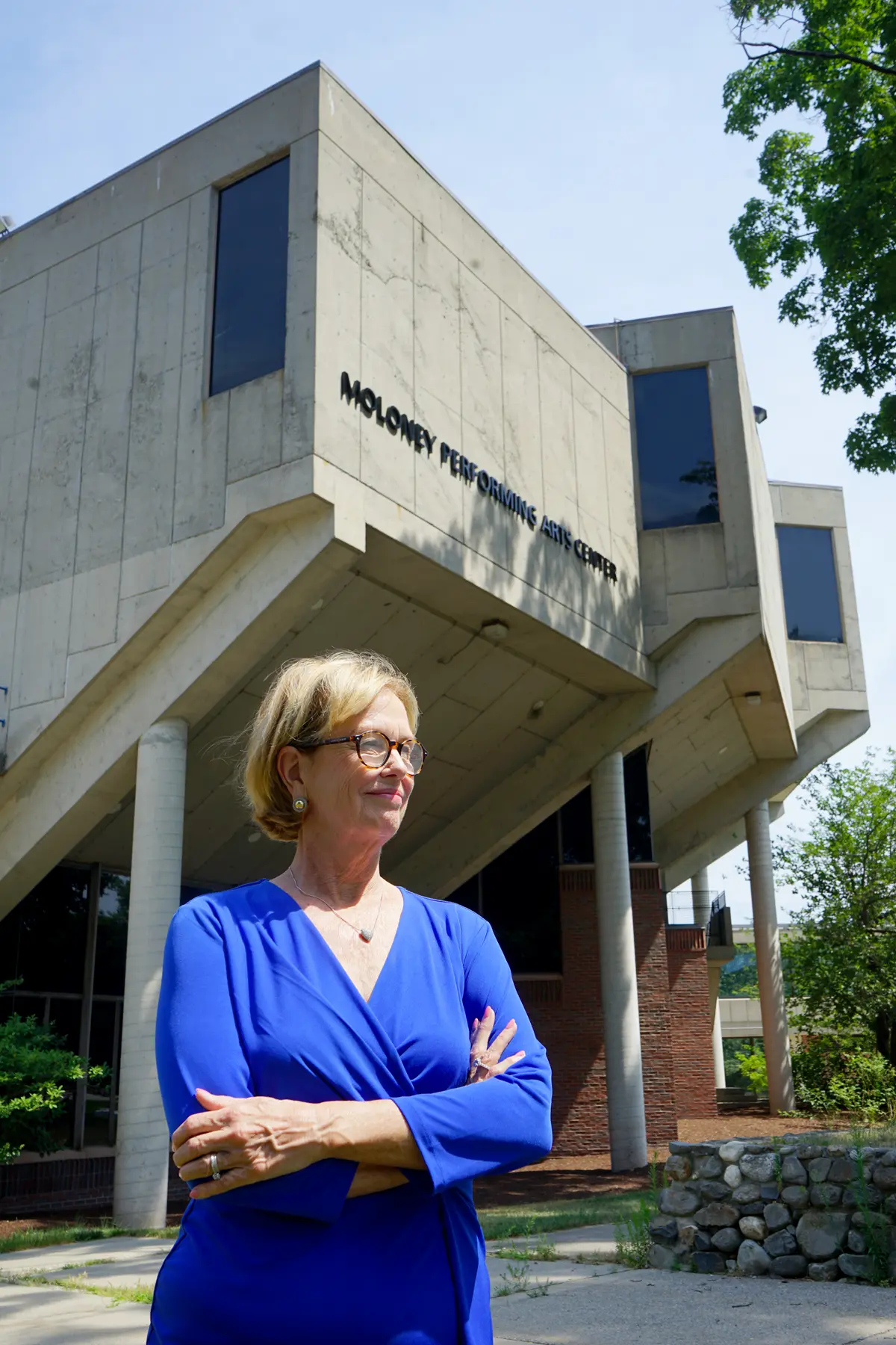 A woman poses with her arms folded standing outside in front of a performing arts center.