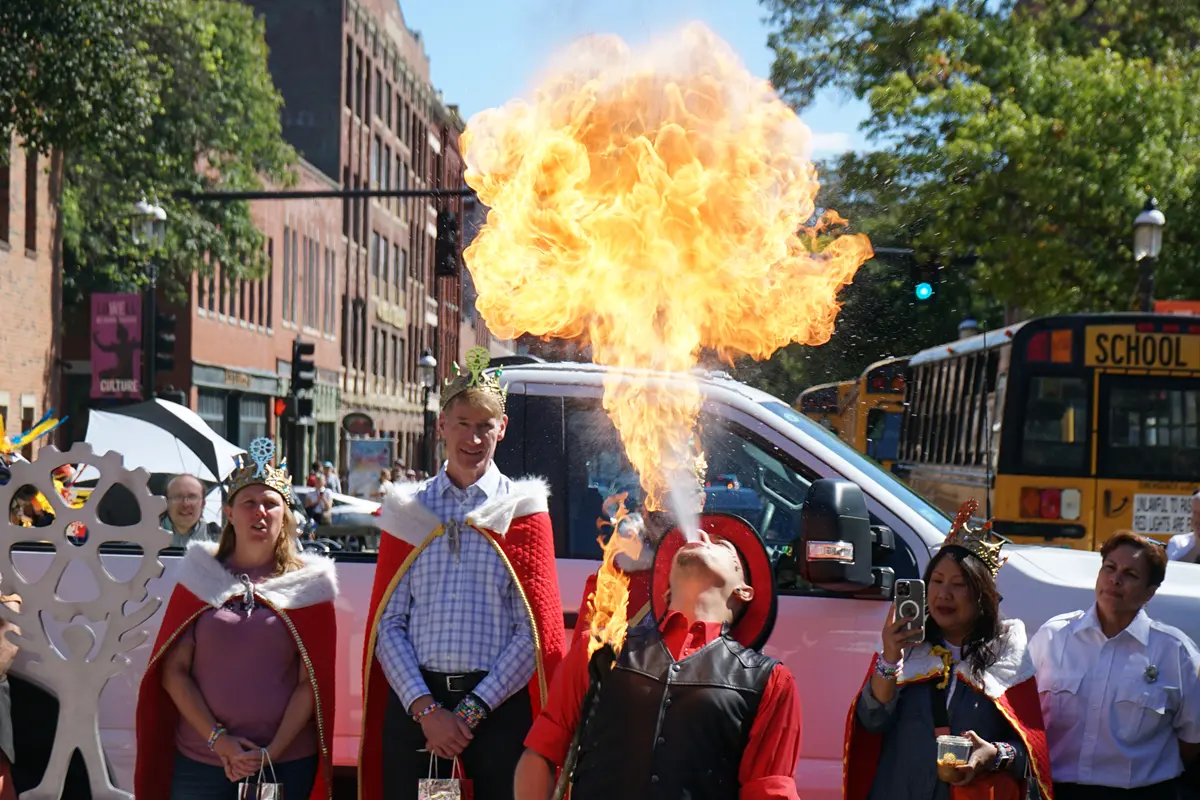 A man breathes fire into the air while people watch in the background while standing in front of a truck.