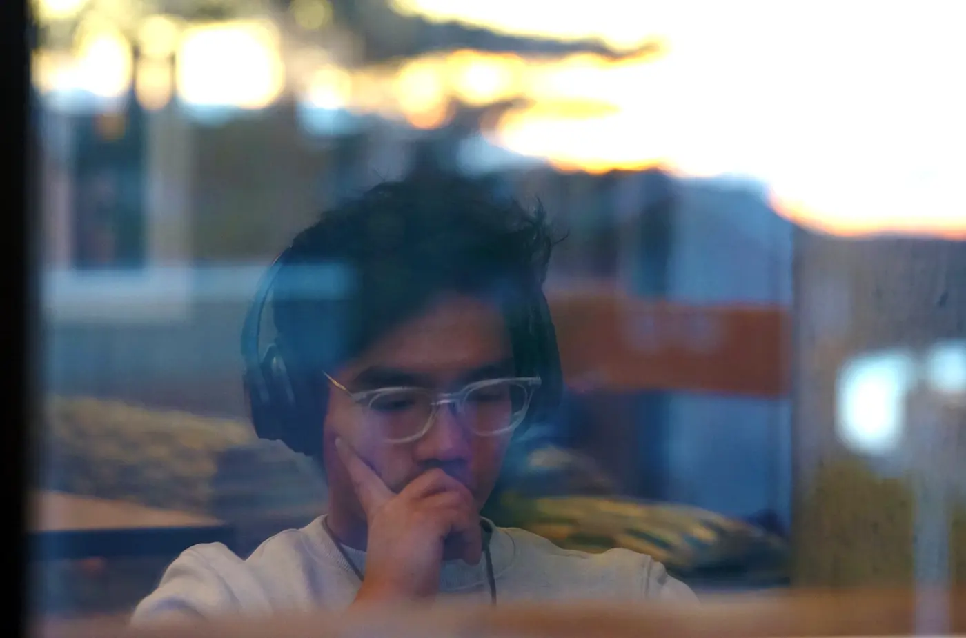 A person with dark hair and glasses listens to headphones while studying in front of a window.