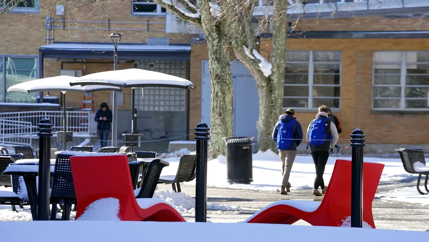 Students wearing backpacks walk amid snow-covered umbrellas with red chairs in foreground