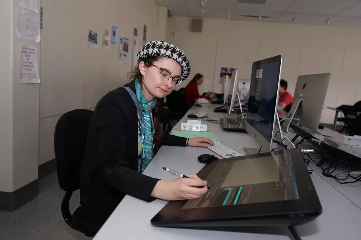A young person stands next to a monitor while speaking to a college art class.