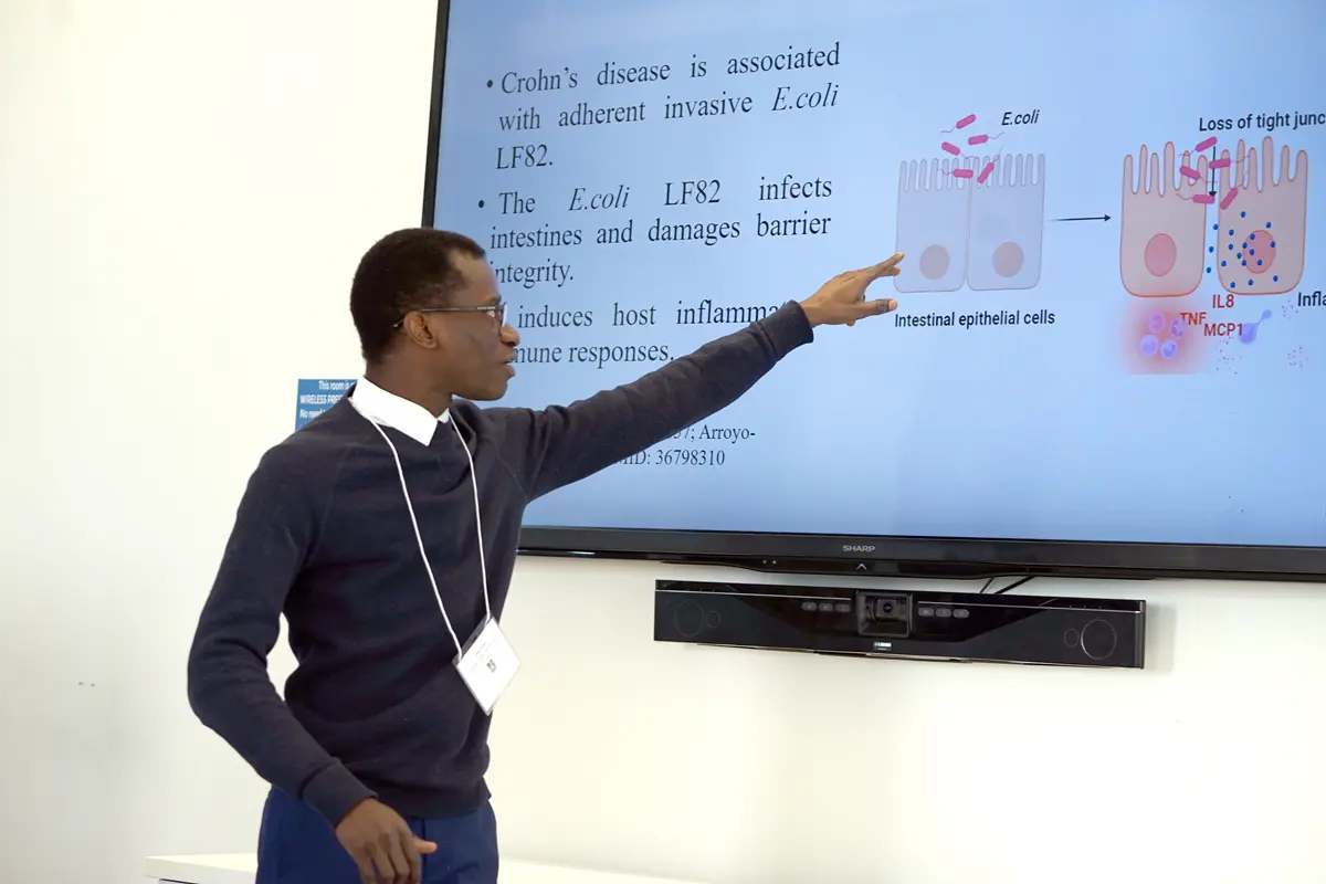 A man in a sweater and collared shirt points to a monitor while giving a presentation.