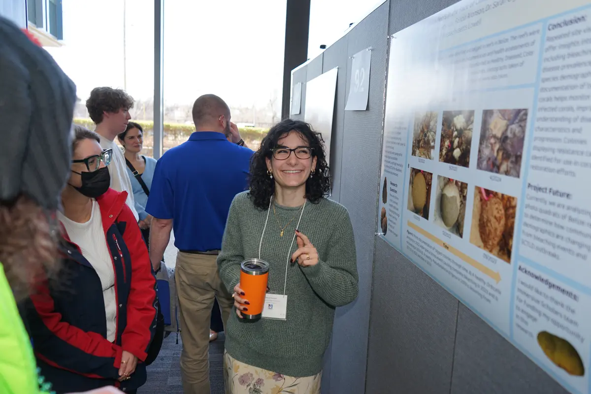 A woman with glasses and holding an orange travel mug smiles while talking about her research poster with a person in a mask.
