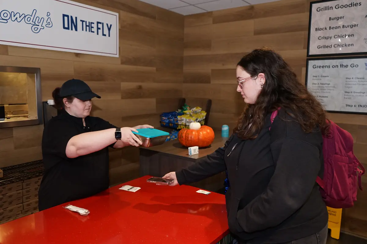 A young woman uses her phone to scan a metal food container that's being held by a woman behind a counter. 
