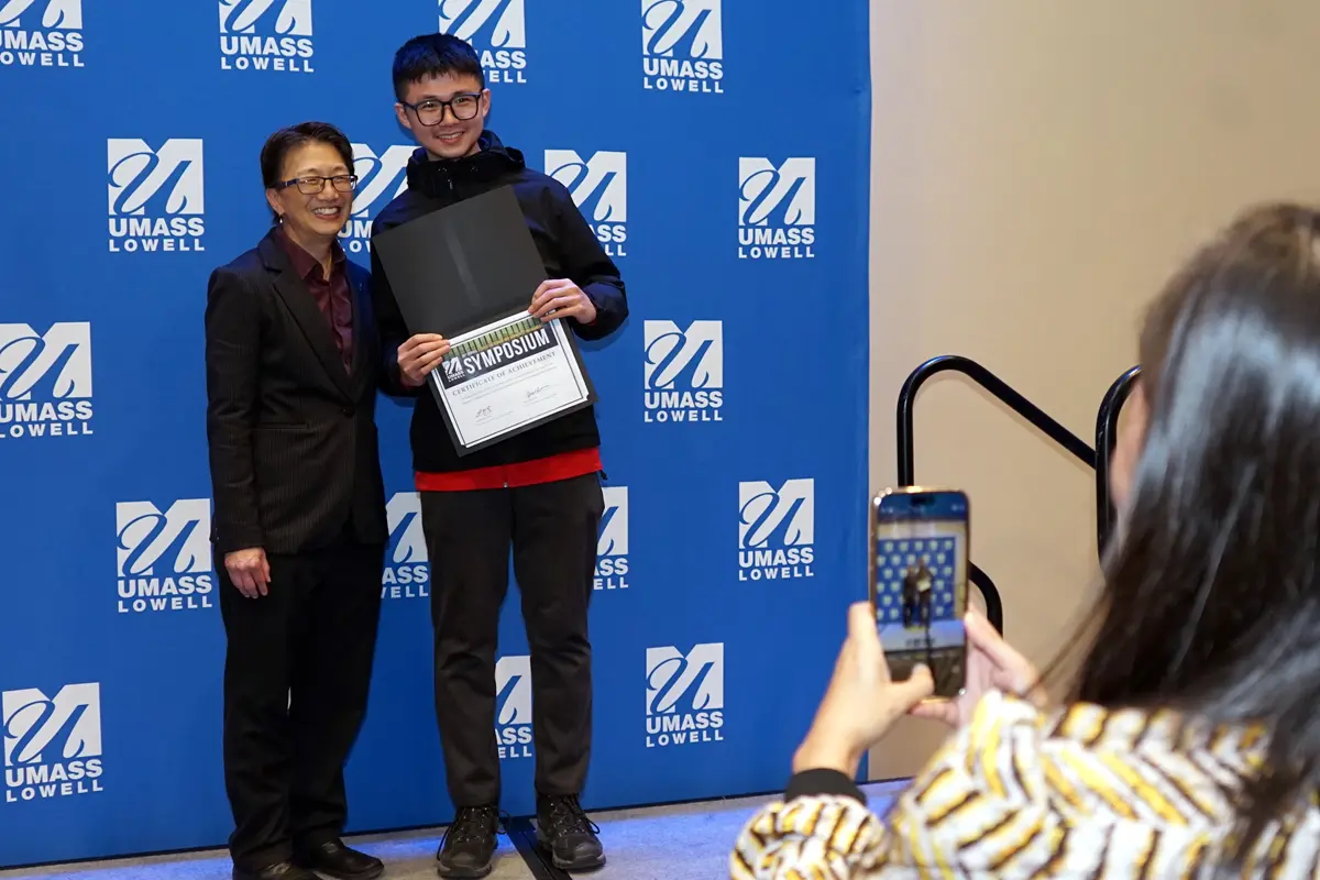 A person take a photo with her smartphone of a young man holding a certificate and posing with a woman on a stage.