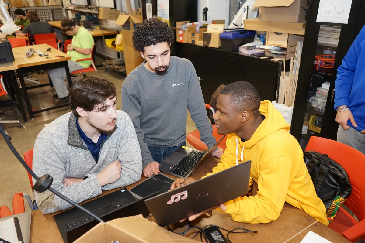 Three young men, two sitting and one standing, work on laptops in a cluttered room.