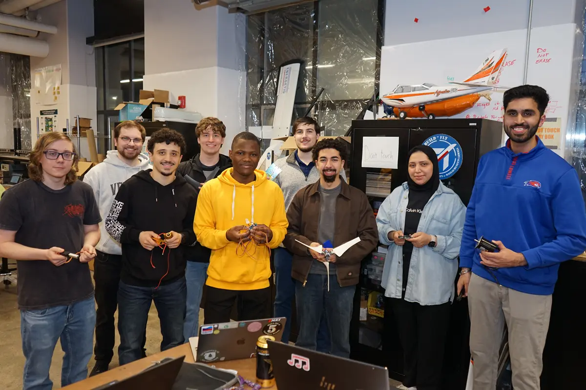 Nine college students pose for a group photo in a workspace. One of them is holding a wind turbine.