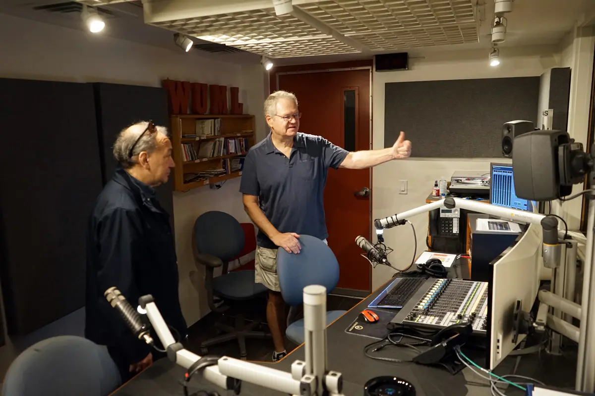 Two men talk while standing in the broadcast studio of a radio station.