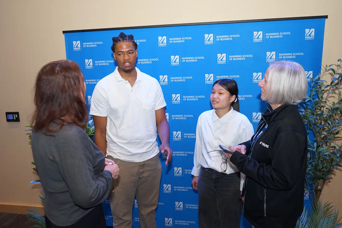 Two college students talk with two women in front of a blue backdrop in a room.
