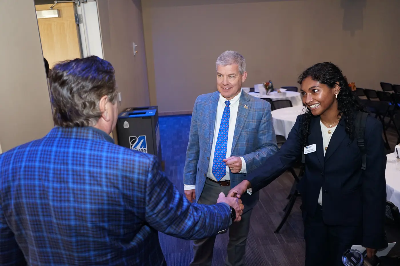 A young woman shakes hands with a man in a checkered blazer while another man looks on.