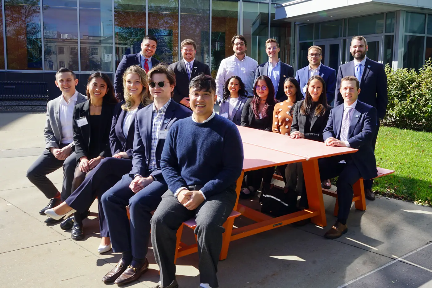 Sixteen men and women pose for a group photo around an orange picnic table in front of a building.