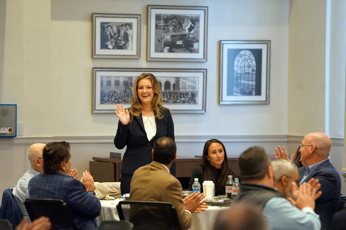 A young woman stands and waves while people sitting at a table applaud.