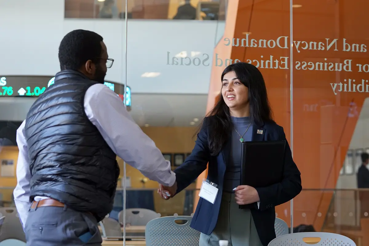 A young woman with dark hair shakes hands with a man wearing a vest in a conference room.
