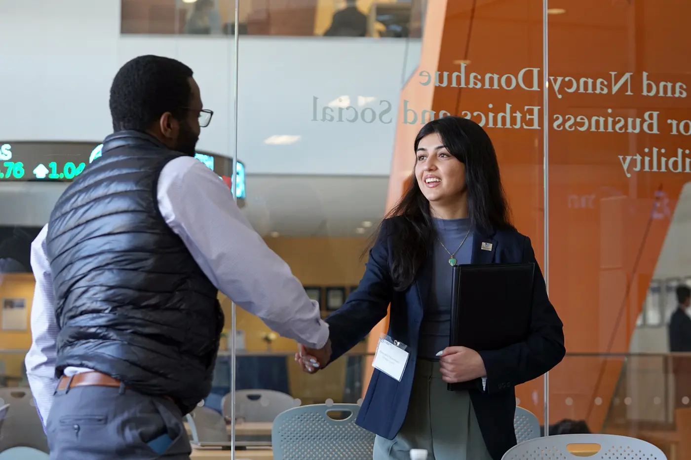 A young woman with dark hair shakes hands with a man wearing a vest in a conference room.